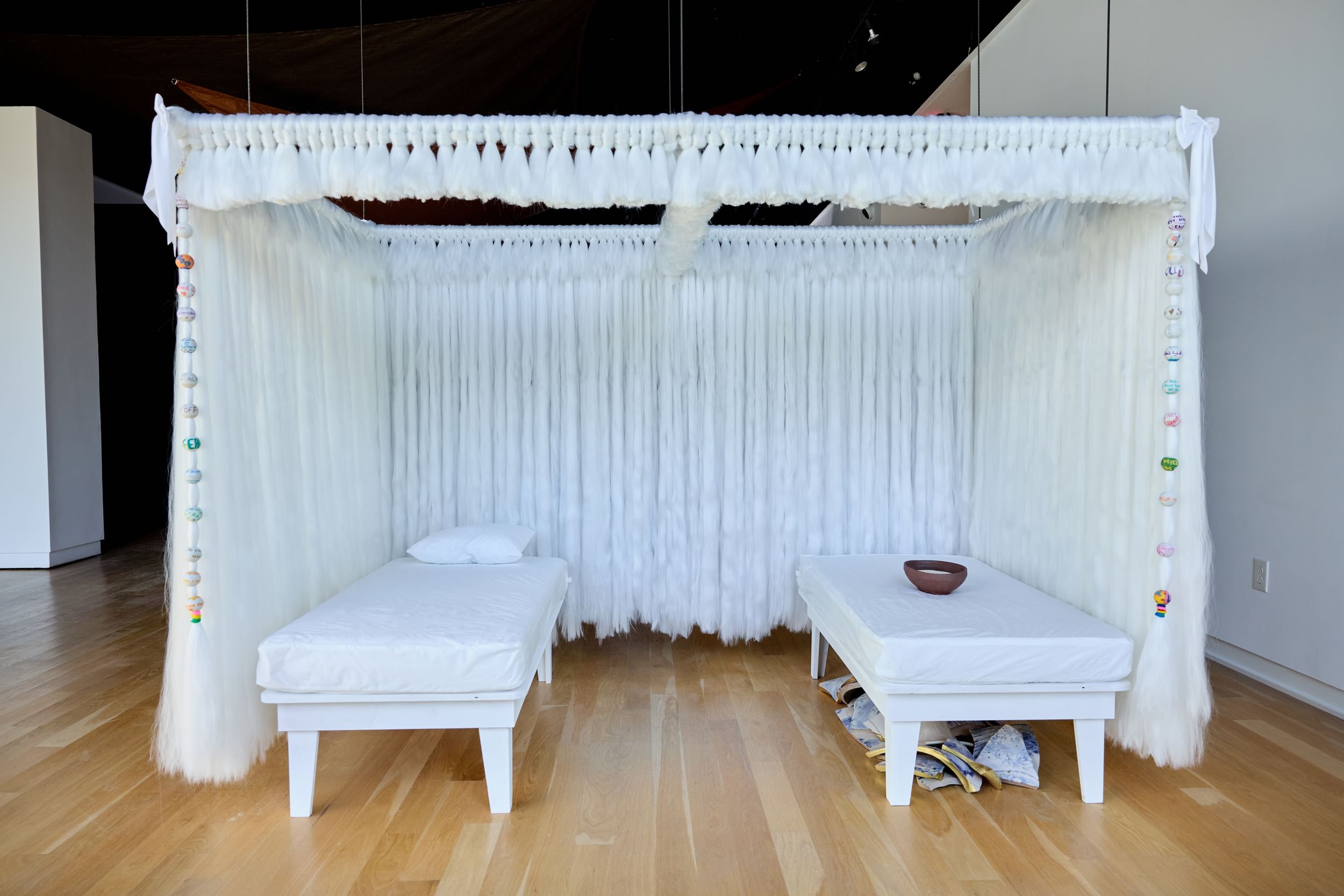 Two white single beds in a small white room, with broken pottery under the one at right, and a brown bowl on top.