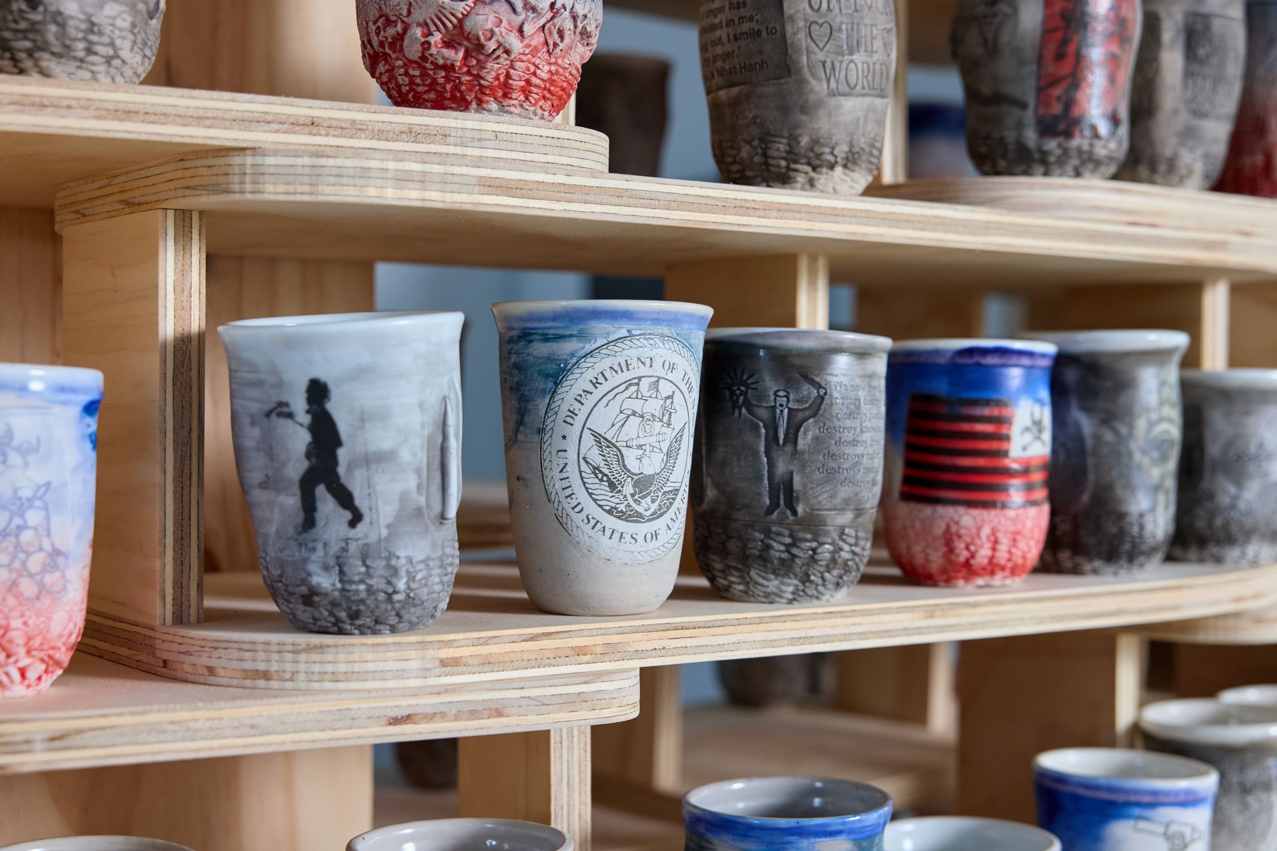 Close-up on wooden shelves full of clay cups with different designs, mostly in reds, blues, and grays.