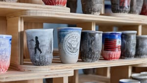 Close-up on wooden shelves full of clay cups with different designs, mostly in reds, blues, and grays.