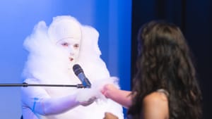 Galligan, seated with a microphone, wears white face paint and a fluffy cotton costume. A woman hands him more cotton.