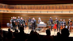 A view from the back of the theater. The audience is in silhouette and 16 onstage musicians stand for their bows.