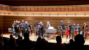 A view from the back of the theater. The audience is in silhouette and 16 onstage musicians stand for their bows.