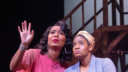 Wilson, in a pink dress, kneels next to Johnson, in a blue dress. They both look outward and Wilson gestures with her hand.