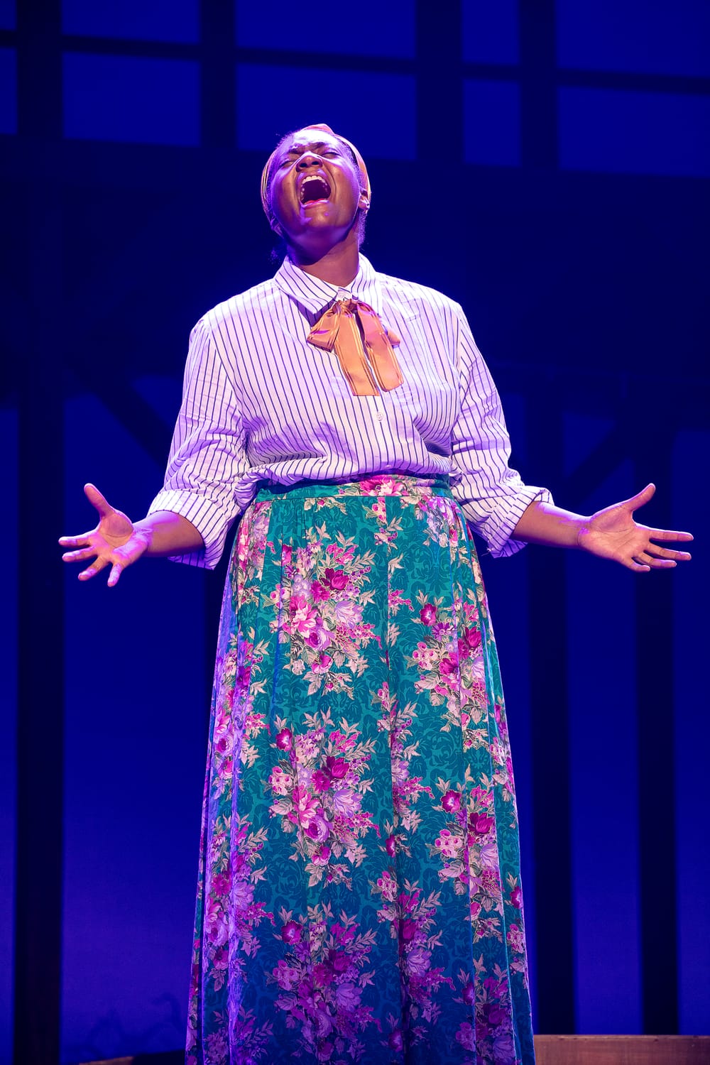 Johnson, a Black woman wearing a long flowered skirt & striped blouse, sings passionately with her hands outspread.