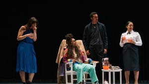 Bowers sits in a white rocking chair and the others group around, looking to the right with different expressions