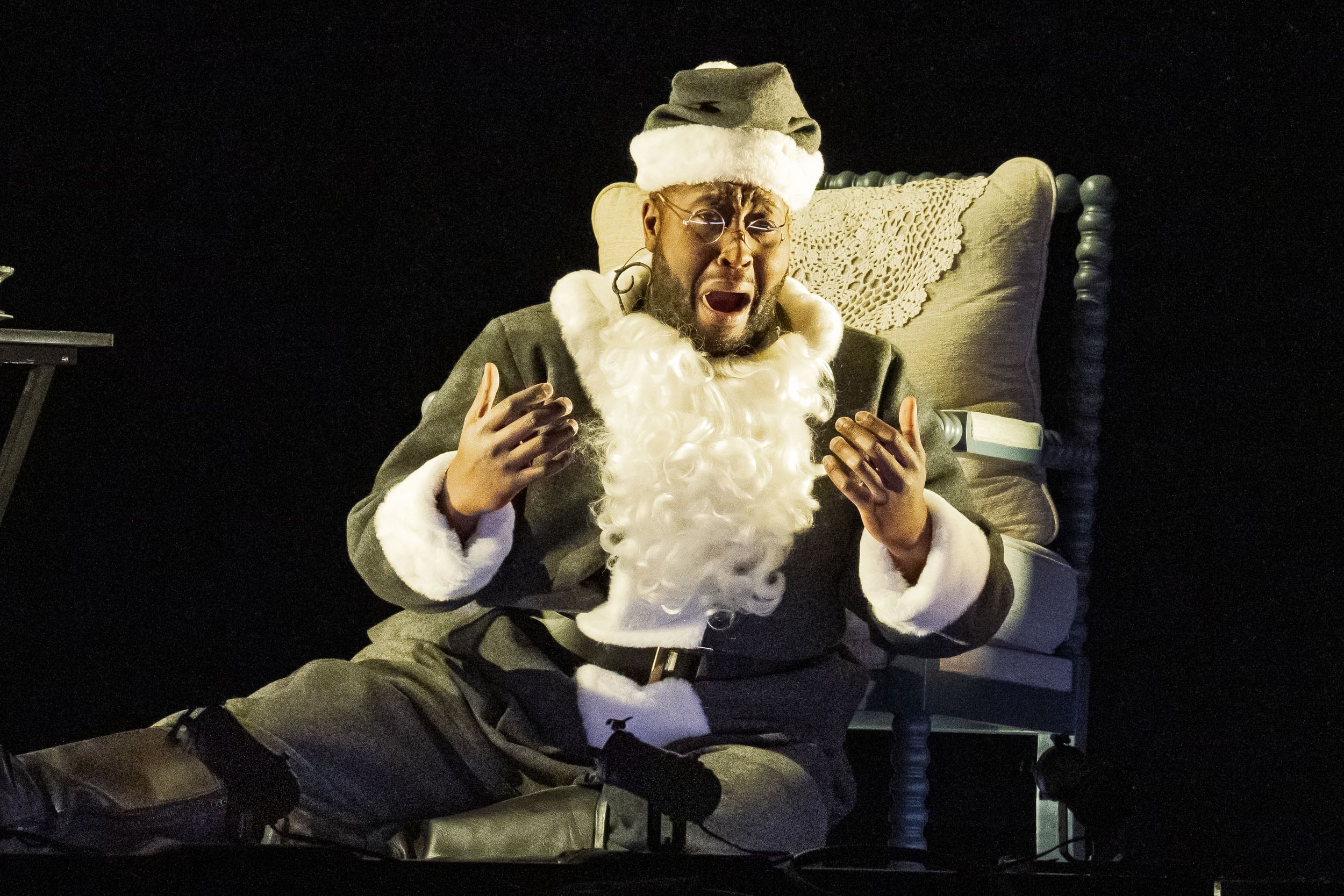 Newton, a Black man wearing a black Santa suit with white trim, sits on the floor and sings with a campy, agonized expression