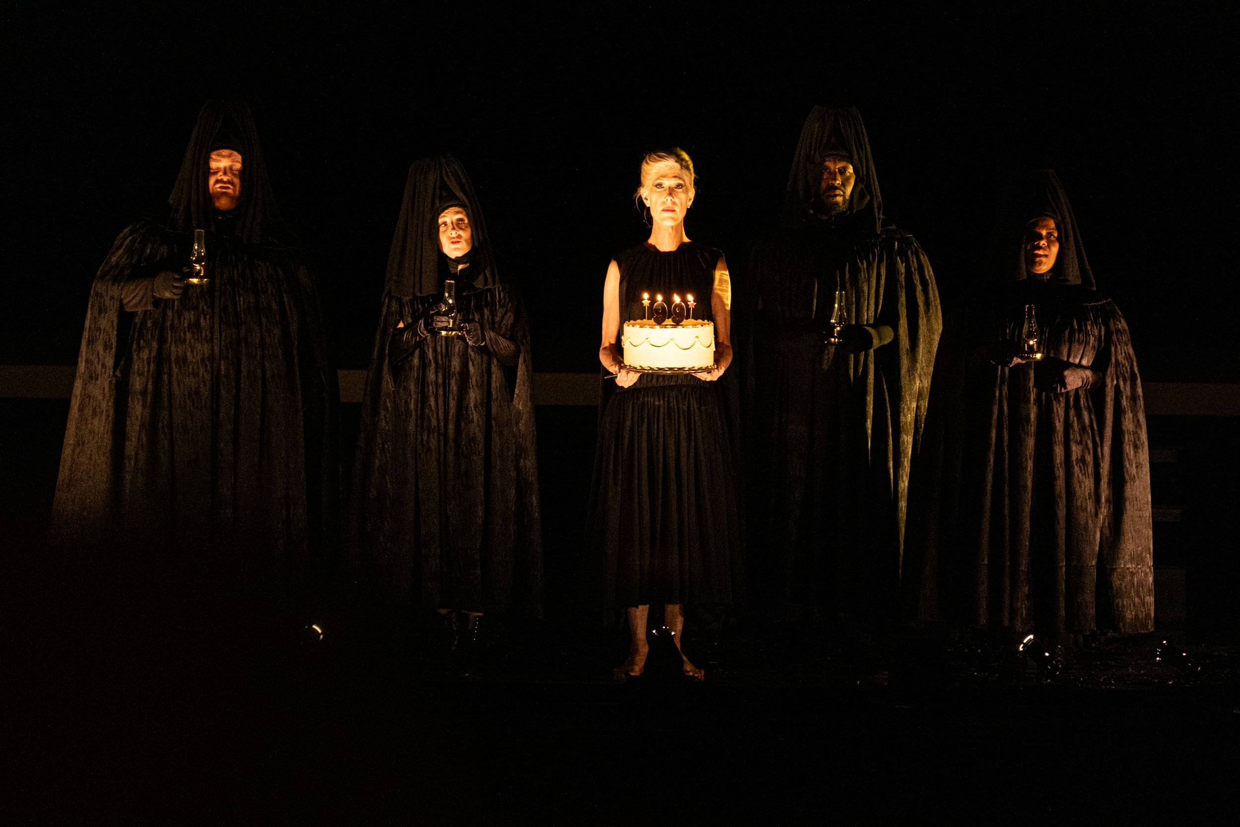 Bond, at center in a black dress holding a birthday cake, with the four singers around her, draped in black costumes.