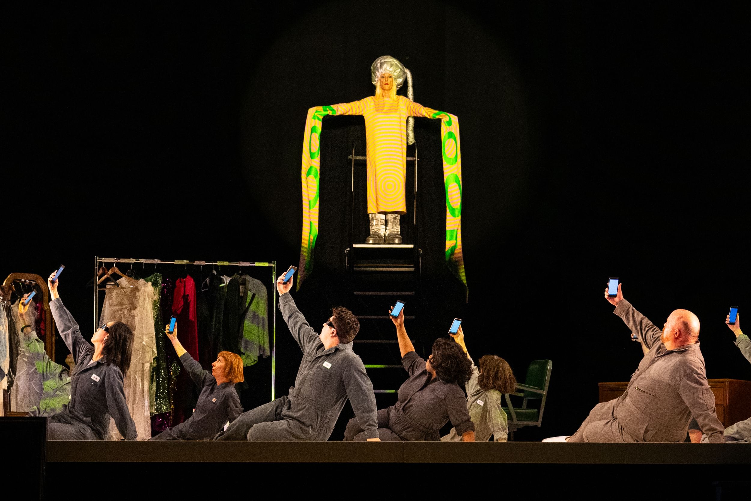 Bond, in yellow with flowing green-lettered sleeves, stands on a platform above 8 cast members in gray suits holding phones