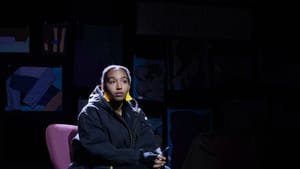 Lutz in character on stage, hands folded, seated. Background is dimly lit, Lutz’s face stern