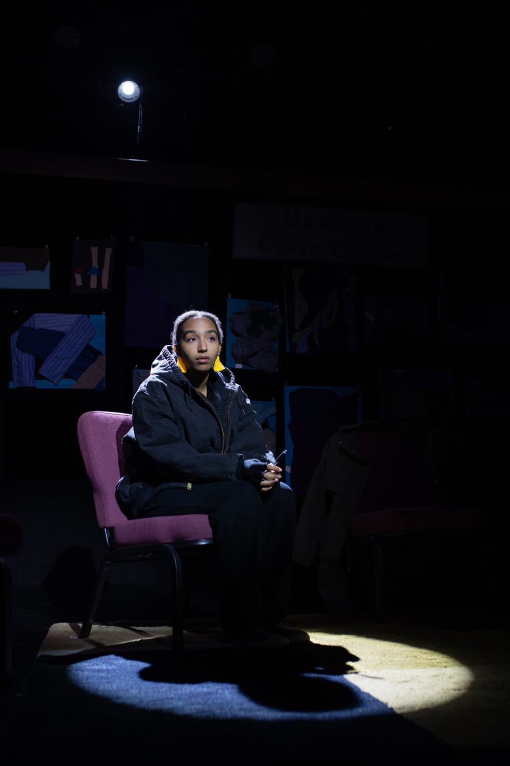 Lutz in character on stage, hands folded, seated. Background is dimly lit, Lutz’s face stern
