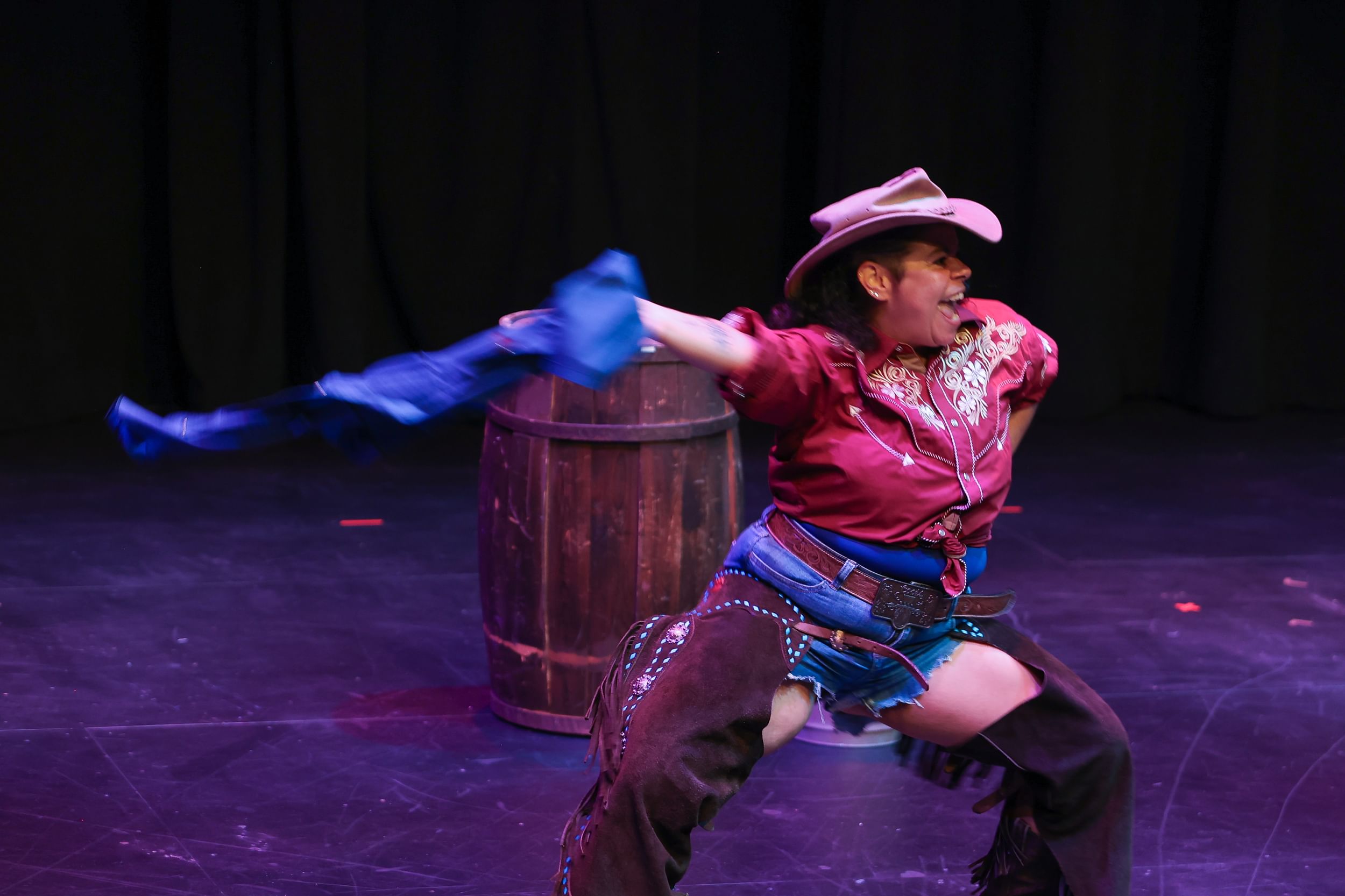 A person in cut off jeans and also shorts, an old Western embroidered shirt, and cowboy hat swings a long cloth like a lasso