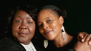 A portrait of Mayson and Wisher, dressed in black with fabric earrings, embrace each other as they look towards the camera