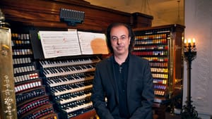 A photo of Conte in a black suit, standing by the organ, posing for the camera. A sheet of music prominent on the organ.