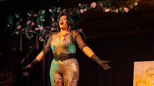 Diorio in drag singing at a standing mic on a stage, wearing a sparkling gold body suit, belt, and boots
