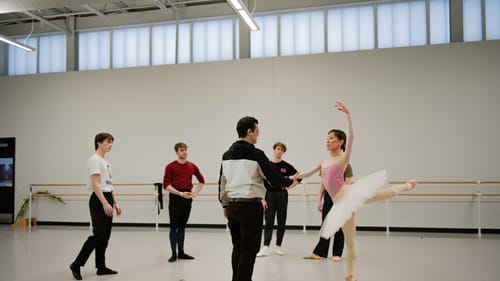 In a white studio, Corella supports Iseda as she poses en point in a tutu while 4 other dancers watch.
