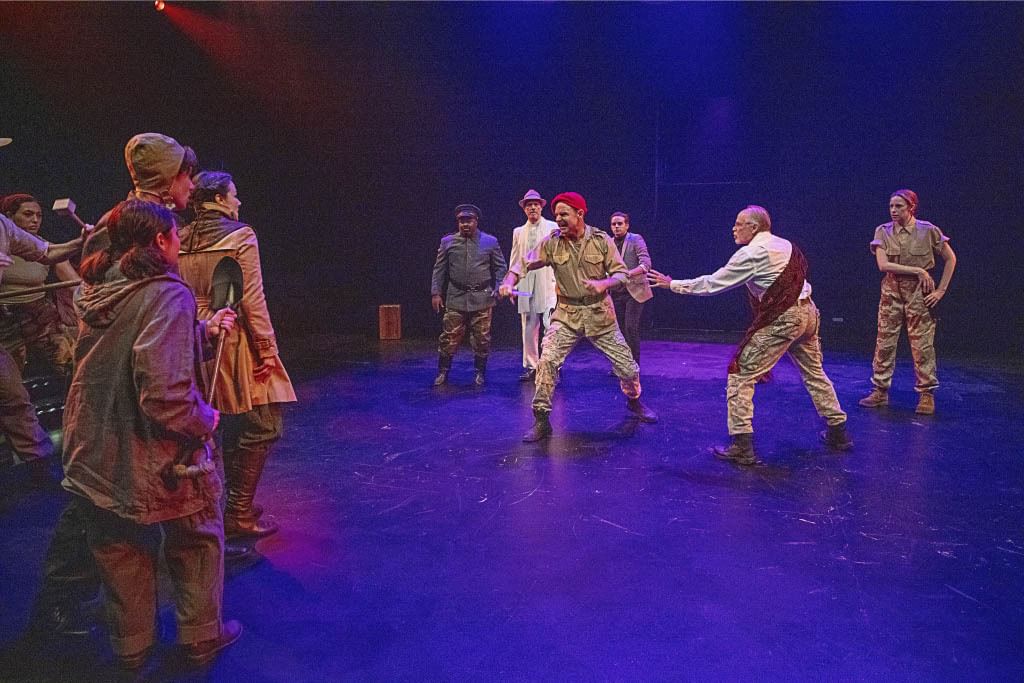 Kent, at center, with military characters holding him back, lunges aggressively at a rabble of commoners at stage left.