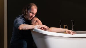 Beschler tenderly inclines his head toward Handler, who lies in a white bathtub, face and one soapy arm visible.