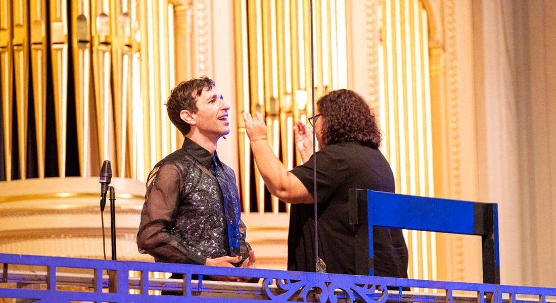 Costanzo sings, to his left is a conductor using their hands, on a stage with bright yellow-gold-ish pillars behind them