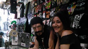 Windfelder and Jolles in all black attire at a store counter look to camera, a wall of apparel lined behind them
