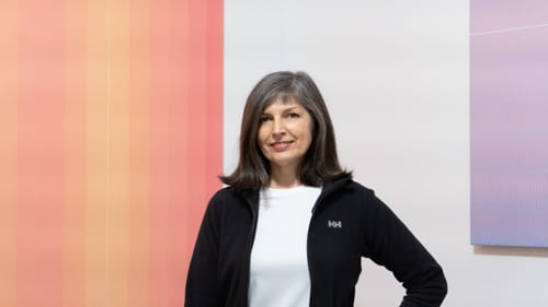 The artist, a white woman with gray-brown hair, poses next to two artworks in the gallery, one in peach tones, one in purple