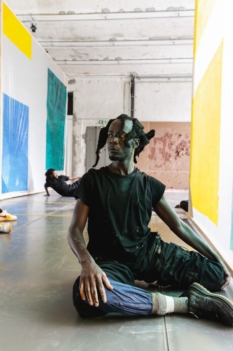 A Black man wearing black, with variously shaped locs, sits on the floor looking warily off to the left.