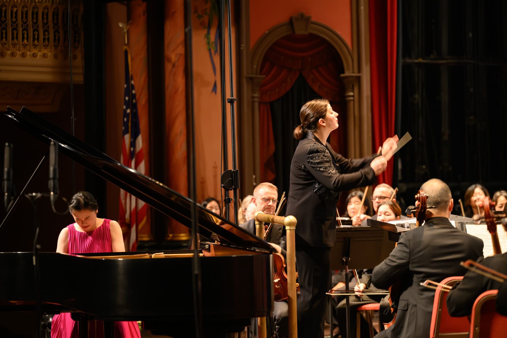 At left, Fei-Fei, in a pink dress, passionately plays the grand piano as Di Russo, in a black blazer, conducts the orchestra