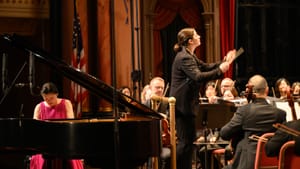 At left, Fei-Fei, in a pink dress, passionately plays the grand piano as Di Russo, in a black blazer, conducts the orchestra