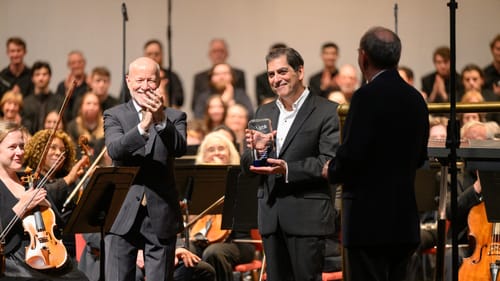 At center, Amado smiles and holds the glass award plaque, surrounded by applauding orchestra leaders and seated musicians.