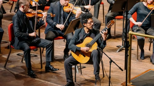 Luiz, a Brazilian guitarist, plays onstage with the orchestra. He wears an elegant black suit.