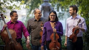 Four players outside with cello and violins—Three laughing looking at each other, with one player smiling towards the camera