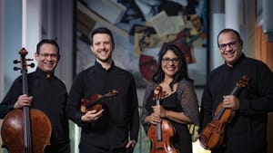 Four players dressed down in mostly all black. Two hold a viola, a cello, and two hold violins