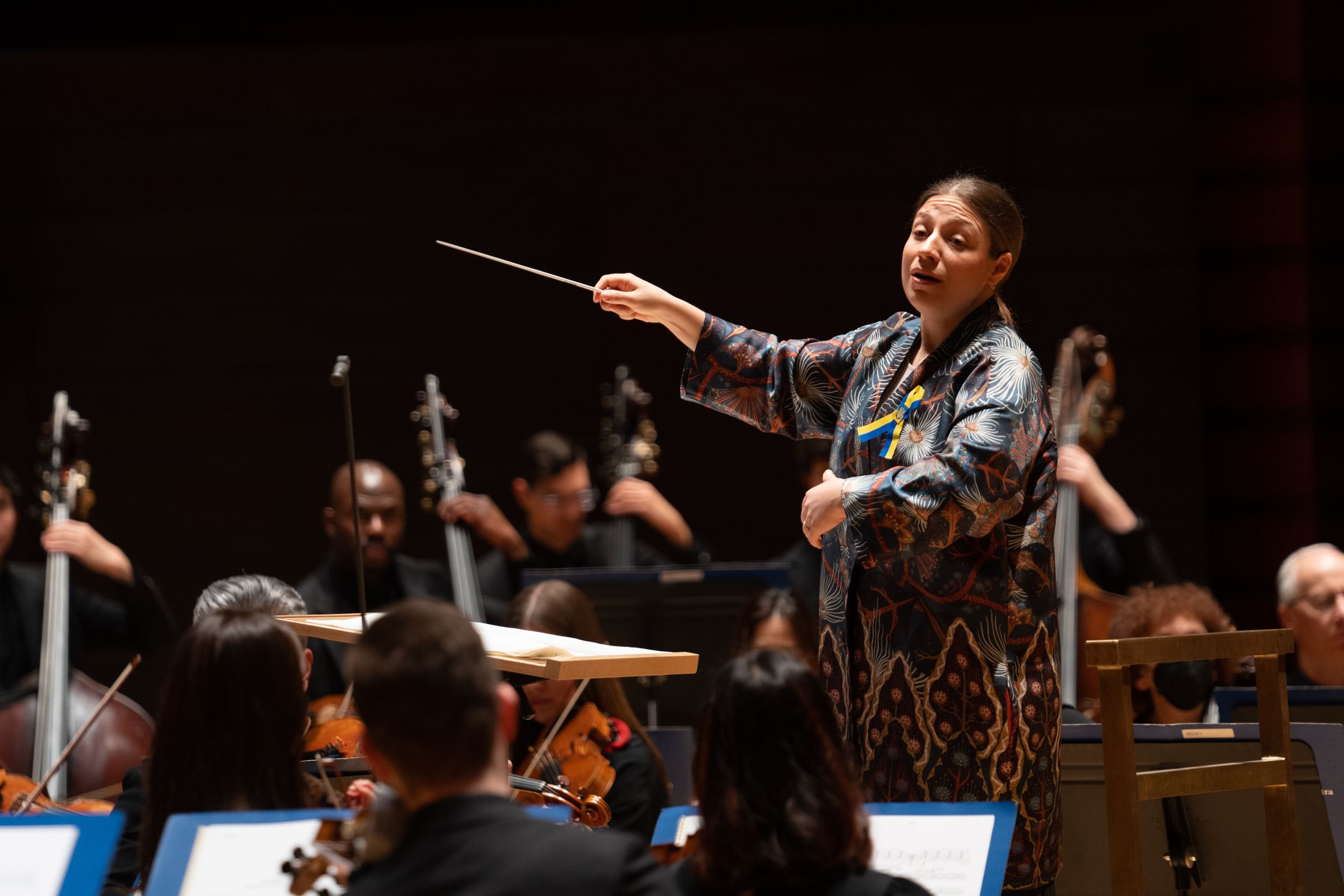 Stasevska, a white woman with brown hair, conducts with one arm extended. She wears a patterned kimono-like shirt