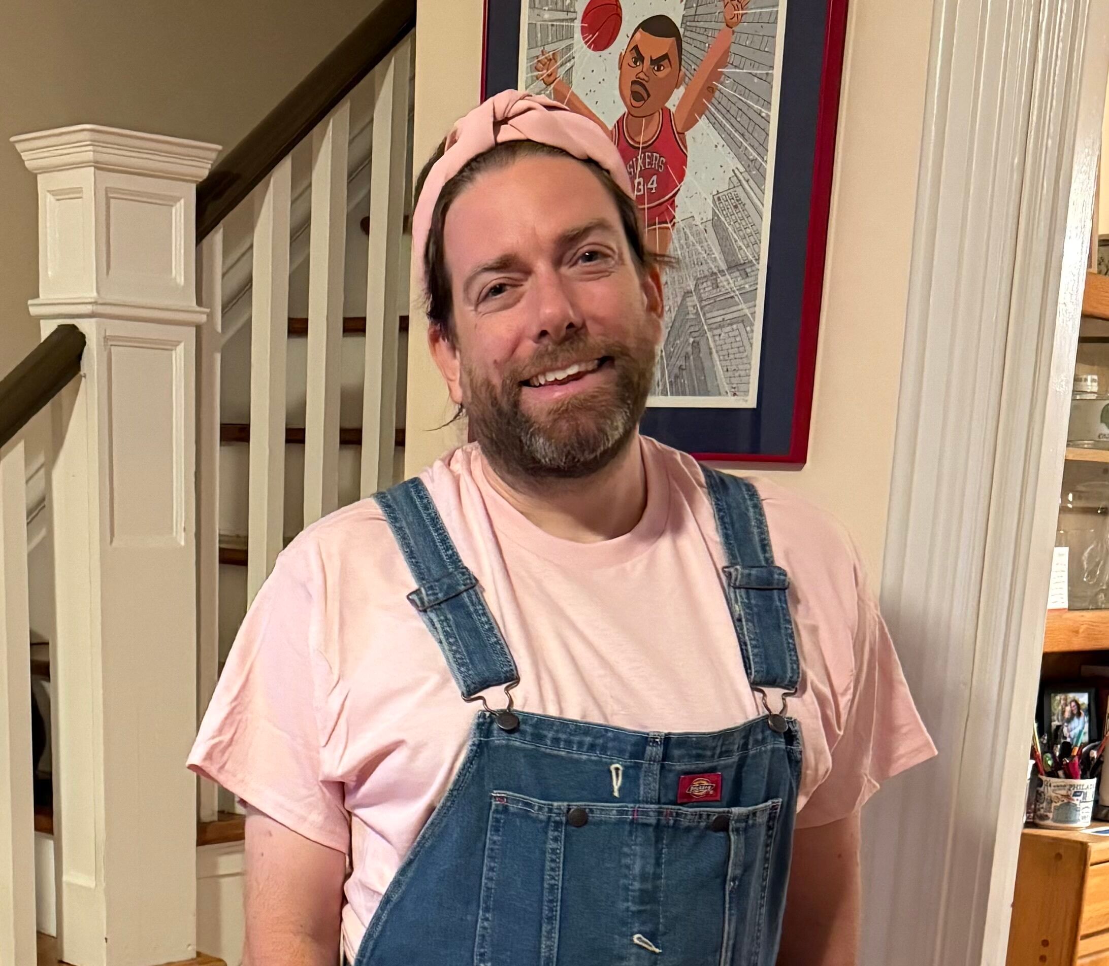 McQuade, a white man with a graying beard, smiles in denim overalls, a pink shirt, and a pink hair wrap.
