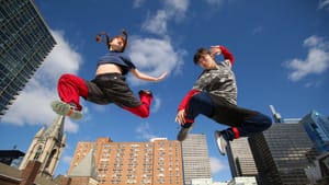 Sood and Smith in mid air in motion, angled looking up, city skyline and a clear blue sky behind them