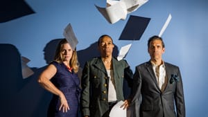 The costumed actors pose in a line with serious faces, X holding papers while others fall through the air around them.