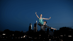 Two ballet dancers in pas de deux, one in all black lifting the other in a leotard, against the Philly skyline at night