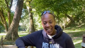 Danie leans on a bridge’s railing during a walk in the green outdoors. They are smiling & wearing purple pants & blue hoodie