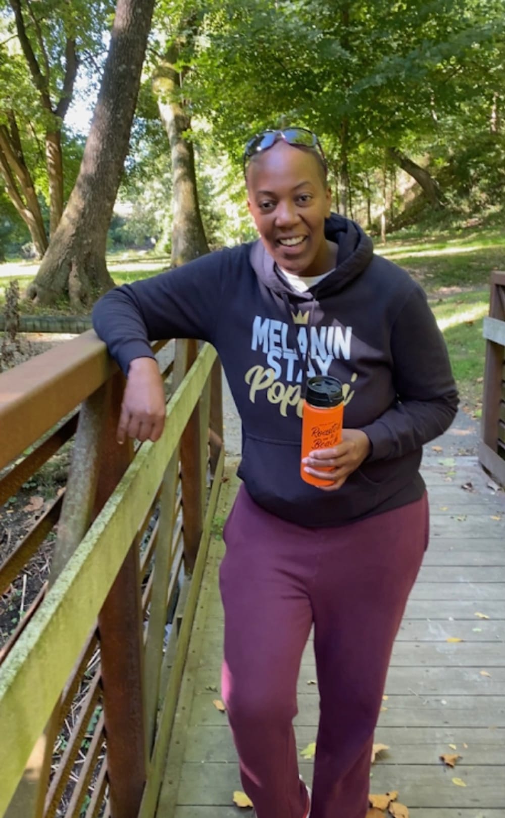 Danie leans on a bridge’s railing during a walk in the green outdoors. They are smiling & wearing purple pants & blue hoodie