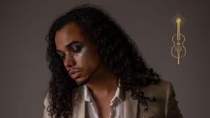 de Jesús in a blazer over a white top that's partly unbuttoned, looks down with eyes closed, posing against a solid backdrop