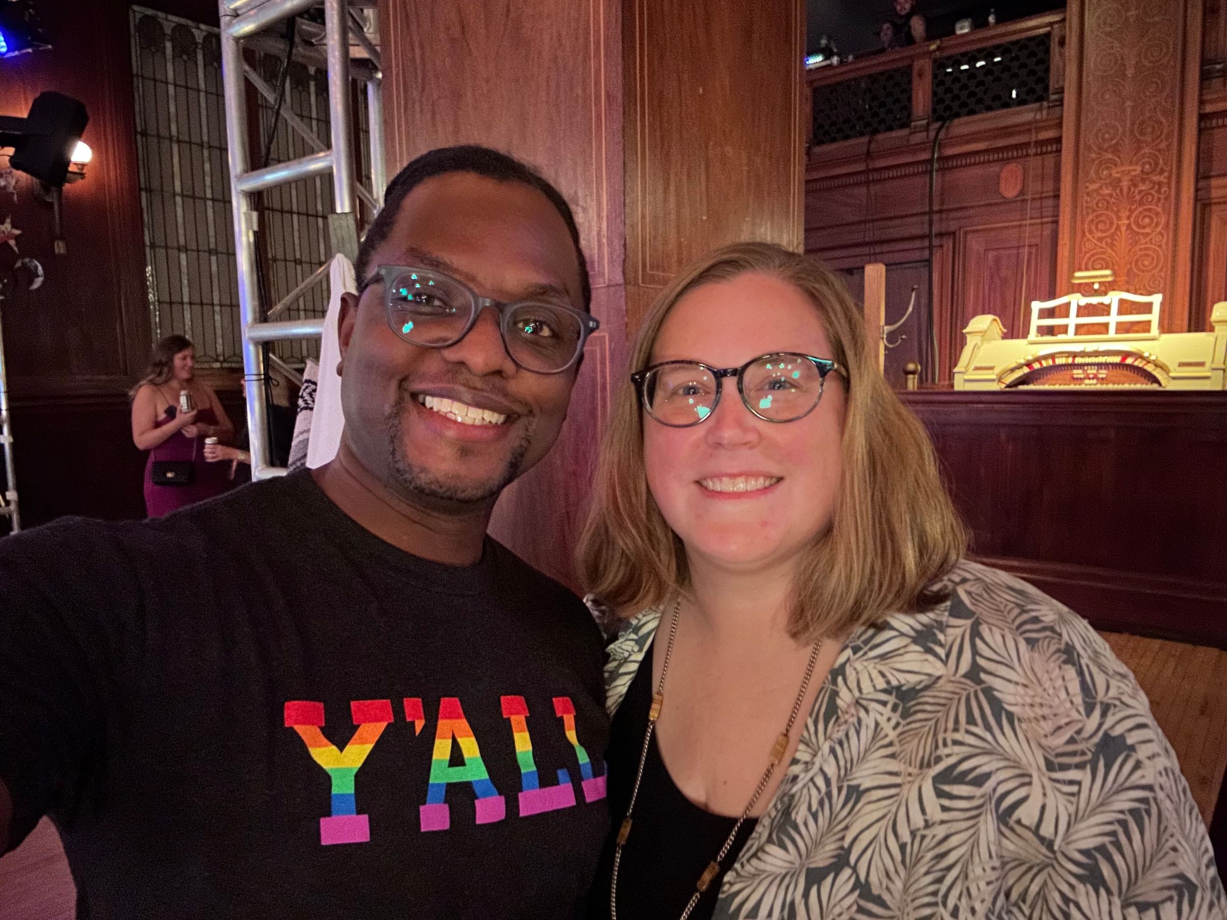 Darnelle, a Black man, poses smiling with Alaina, a white woman, in a small wood-paneled ballroom.