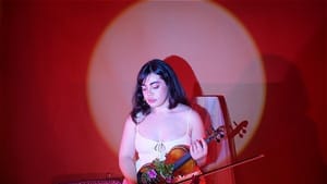 A woman sits holding a violin, lit by soft red light. She wears white, with flowers, books, other artifacts around her.