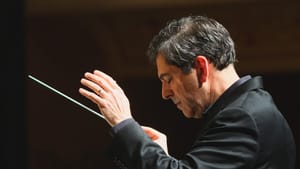 Amado in a black formal jacket, head down, lifting a conductor baton in one hand, in a music hall.