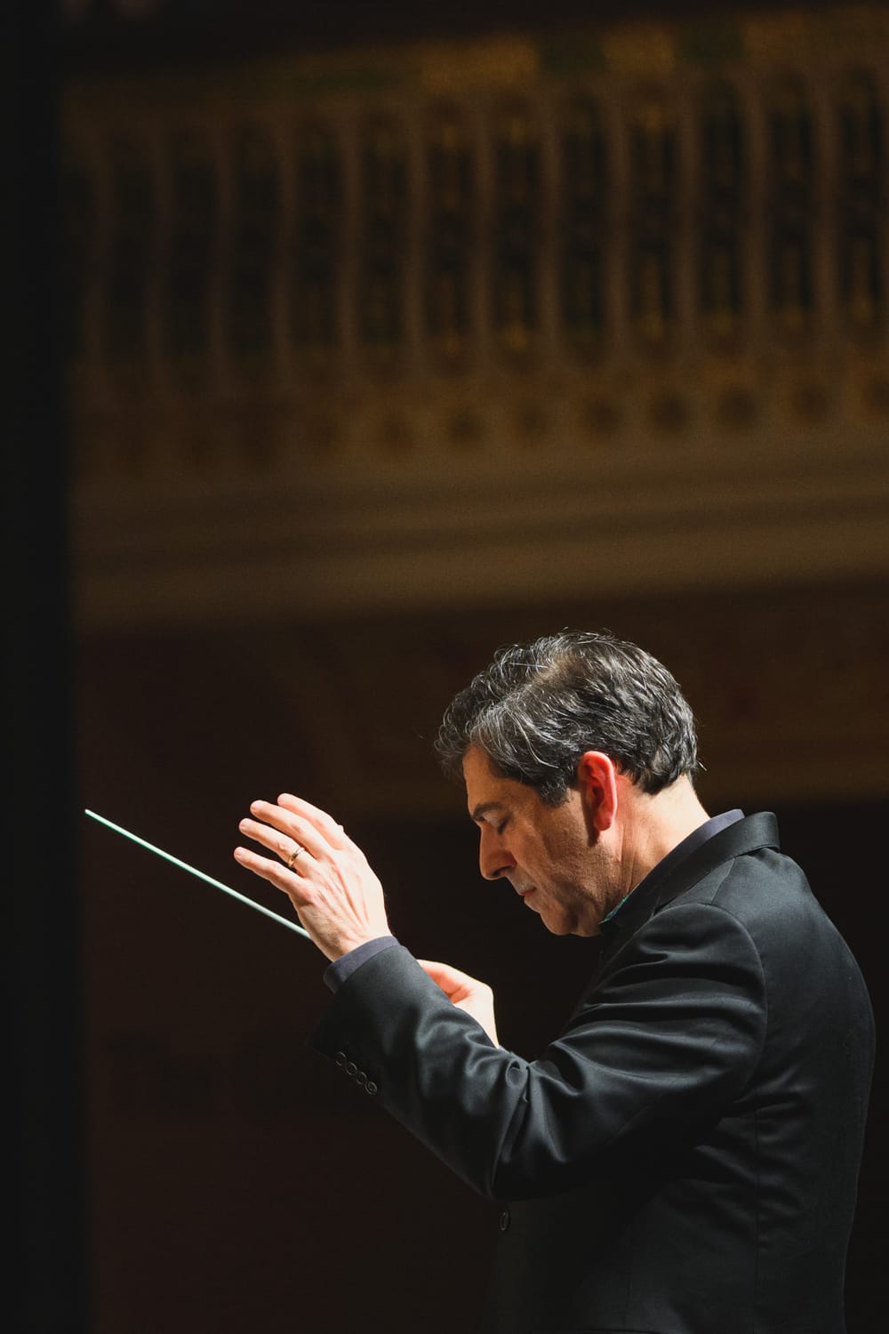 Amado in a black formal jacket, head down, lifting a conductor baton in one hand, in a music hall.