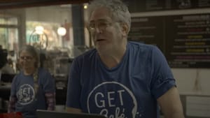 David, a white man with glasses and short gray hair, works behind the register in a coffee shop. He wears a blue t-shirt.