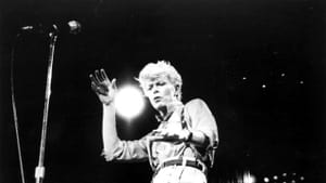 A black and white photo of David Bowie performing on stage, dancing. The background is dark, a mic stands in front of him