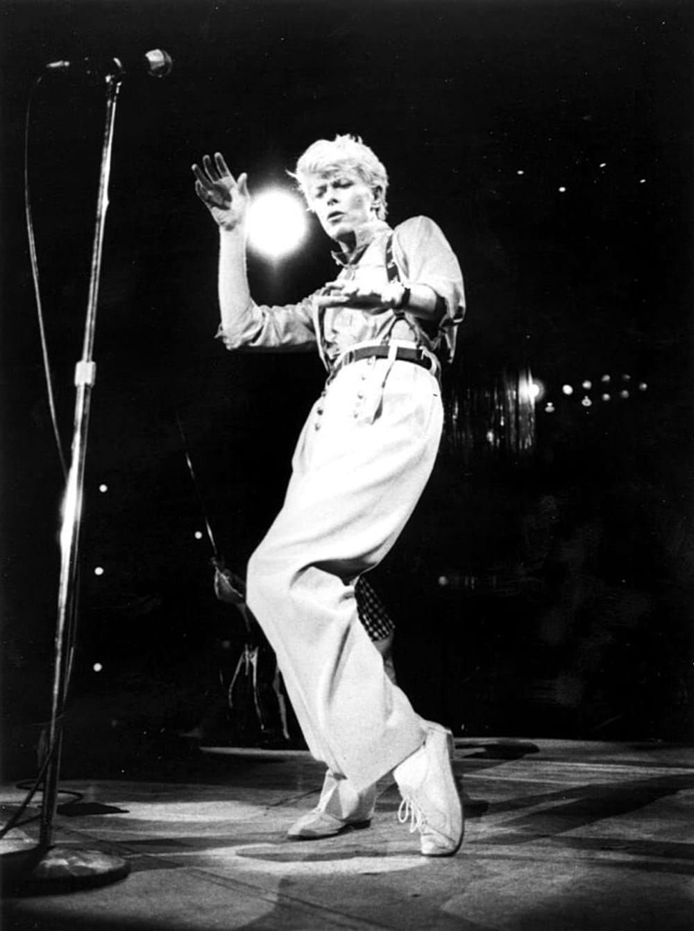 A black and white photo of David Bowie performing on stage, dancing. The background is dark, a mic stands in front of him