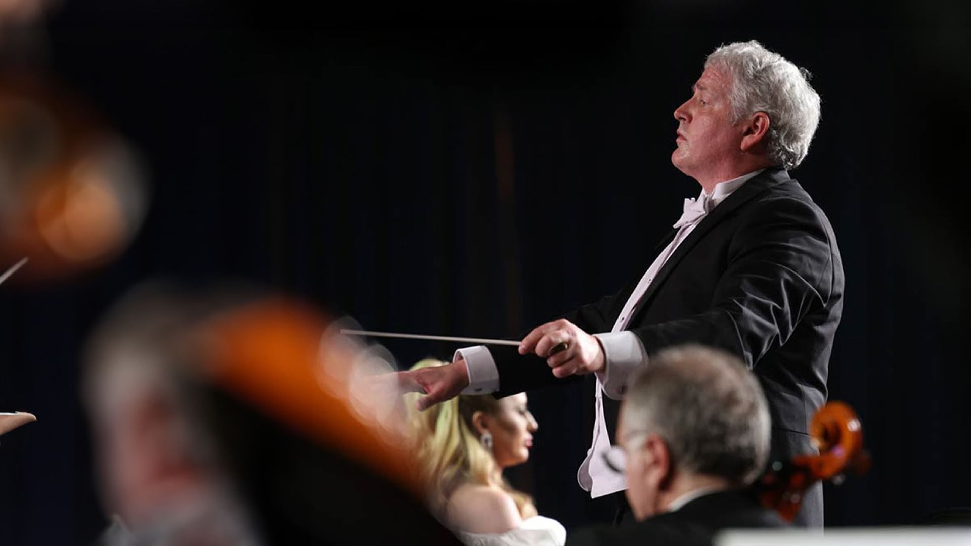 Close up side view of Hayes in classic formalwear, black jacket, white shirt and tie, conducts among musicians