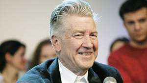 A medium close up portrait of Lynch, with blonde/gray hair, a black jacket, in front of a mic, smiling or laughing