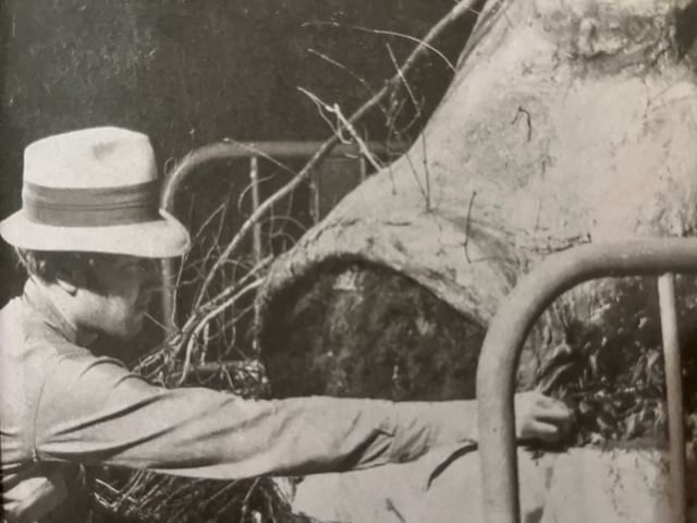 Black & white vintage photo of young Lynch, in profile, wearing a hat, touching a surreal set piece surrounded by sticks