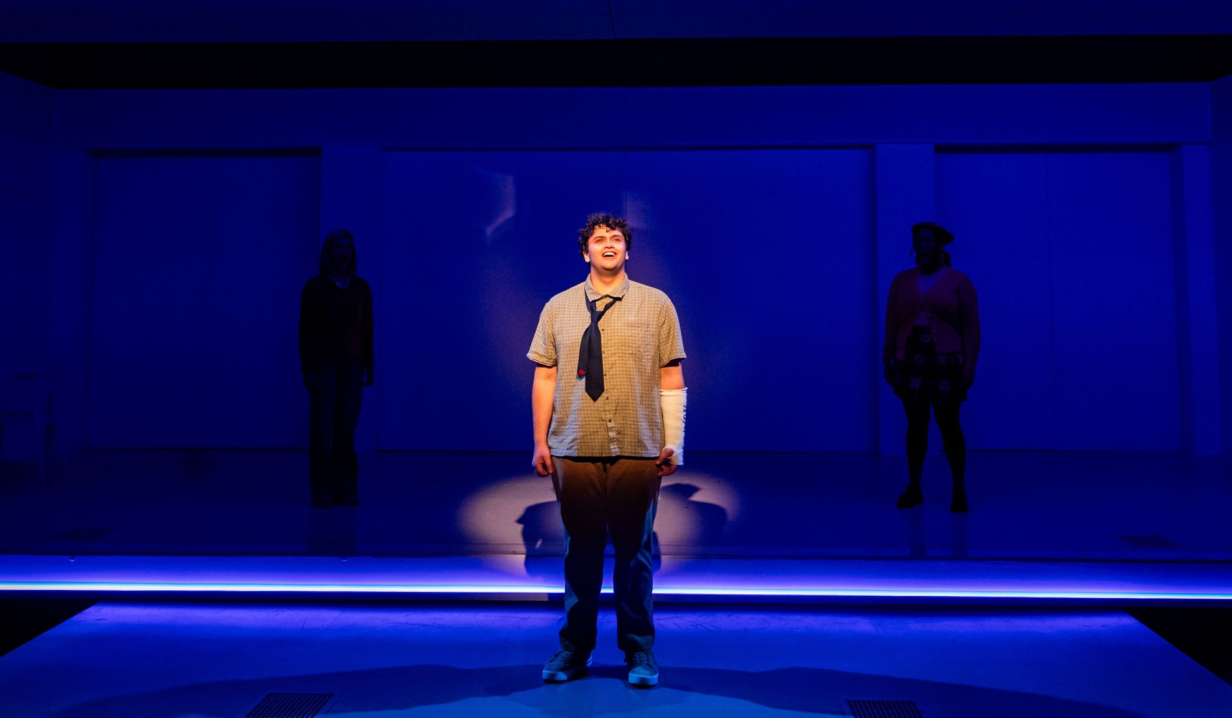 Kaushesh, with curly mop of hair, plaid short-sleeve button-down & short tie, cast on his left arm, sings on a blue-lit stage